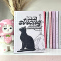 Notebook with a black cat illustration and 'Be strong and courageous' text, surrounded by other books and a decorative figure.