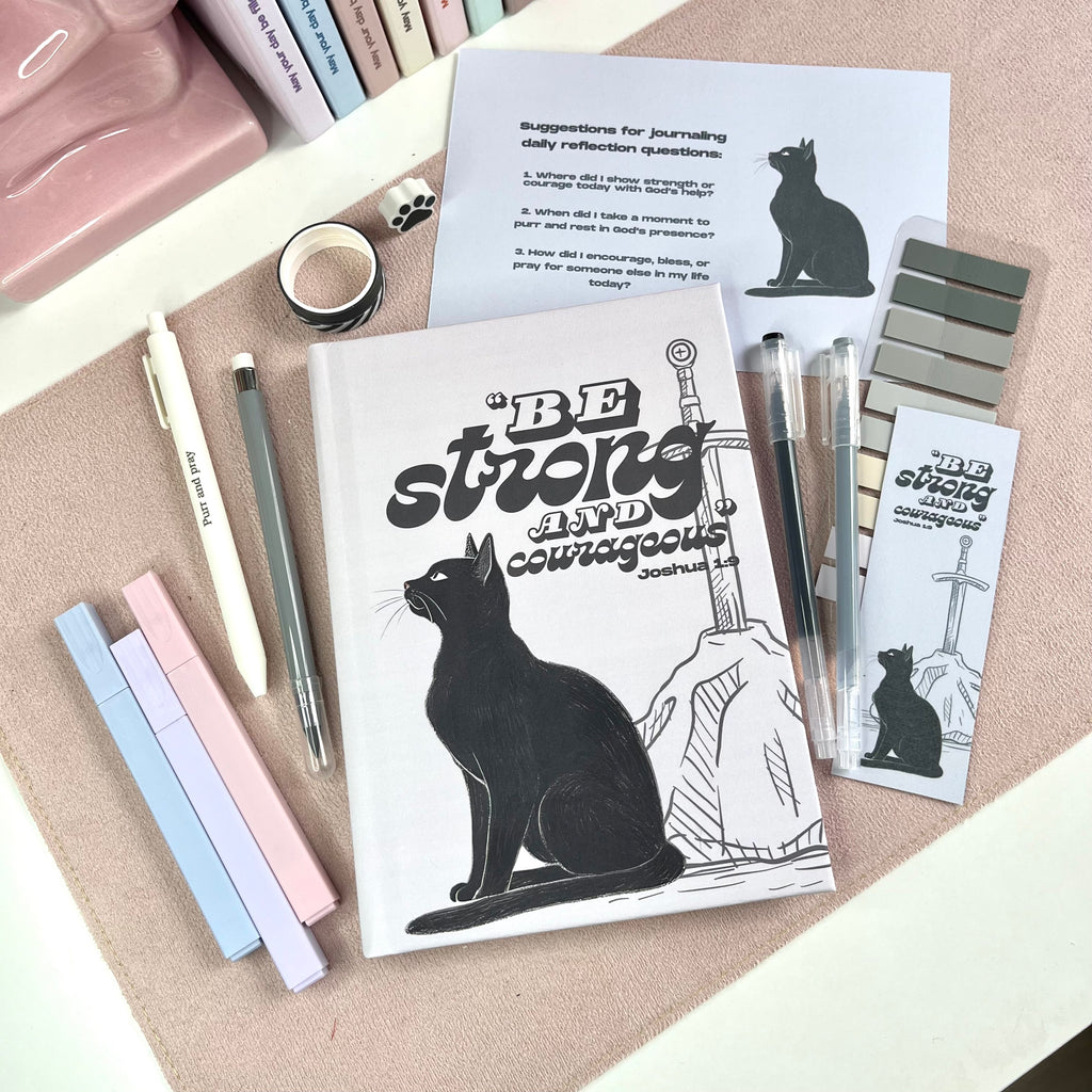 Notebook with a black cat illustration and motivational text, surrounded by pens and stationery items on a textured surface.