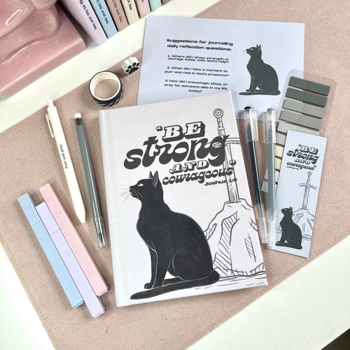 Notebook with a black cat illustration and motivational text, surrounded by pens and stationery items on a textured surface.