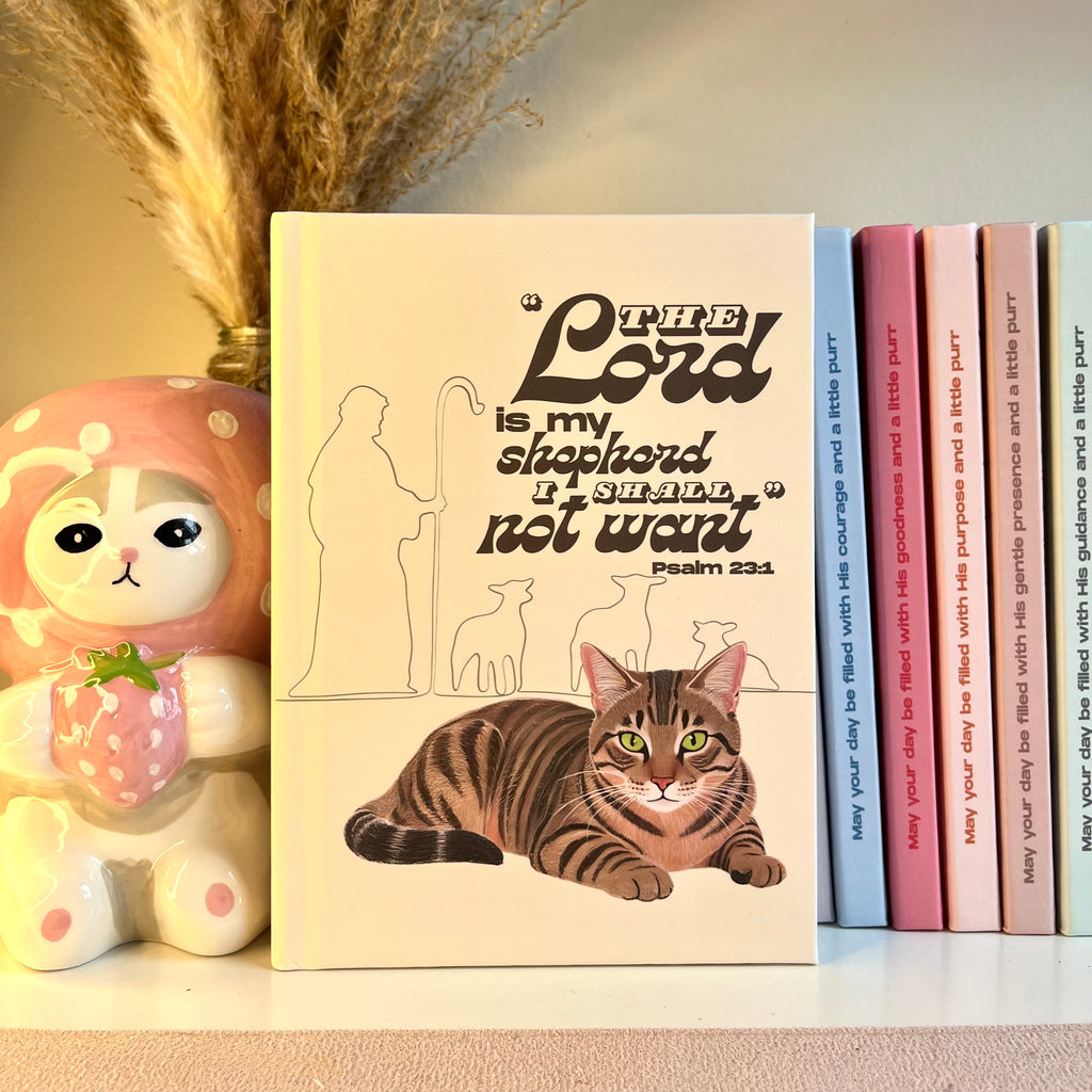 Yellow notebook with a brown tabby cat illustration and Psalm 23:1 text on a shelf with books and a decorative item.