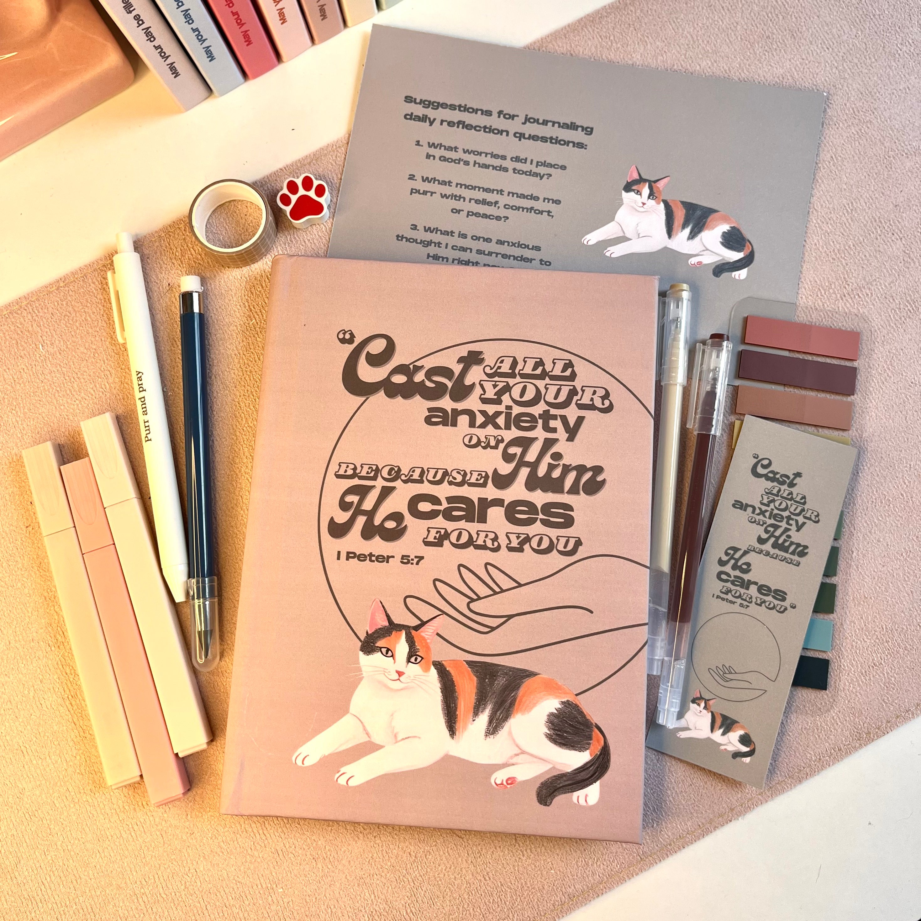 Notebook with calico cat illustration and motivational quote, surrounded by stationery items on a textured surface.