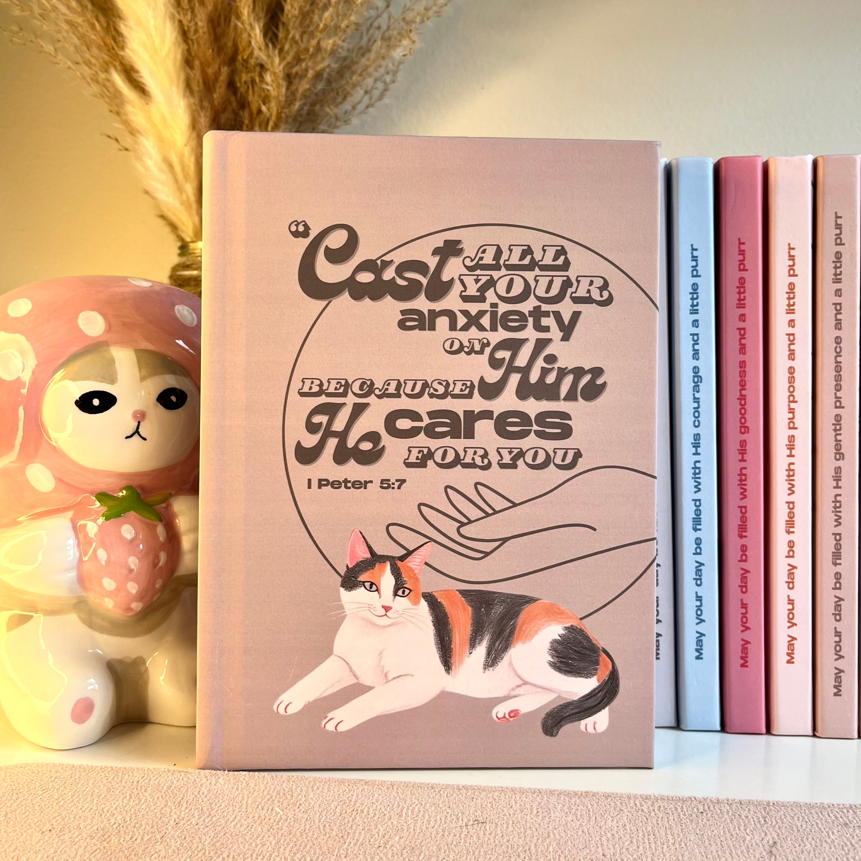 Brown notebook with a calico cat illustration and motivational Bible quote on a shelf with decorative items.