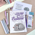 Notebook with a gray cat illustration and text, surrounded by pens and bookmarks on a textured surface.