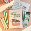 Notebook with gray tabby cat illustration and motivational quote, surrounded by pens and stationery items on a wooden surface.