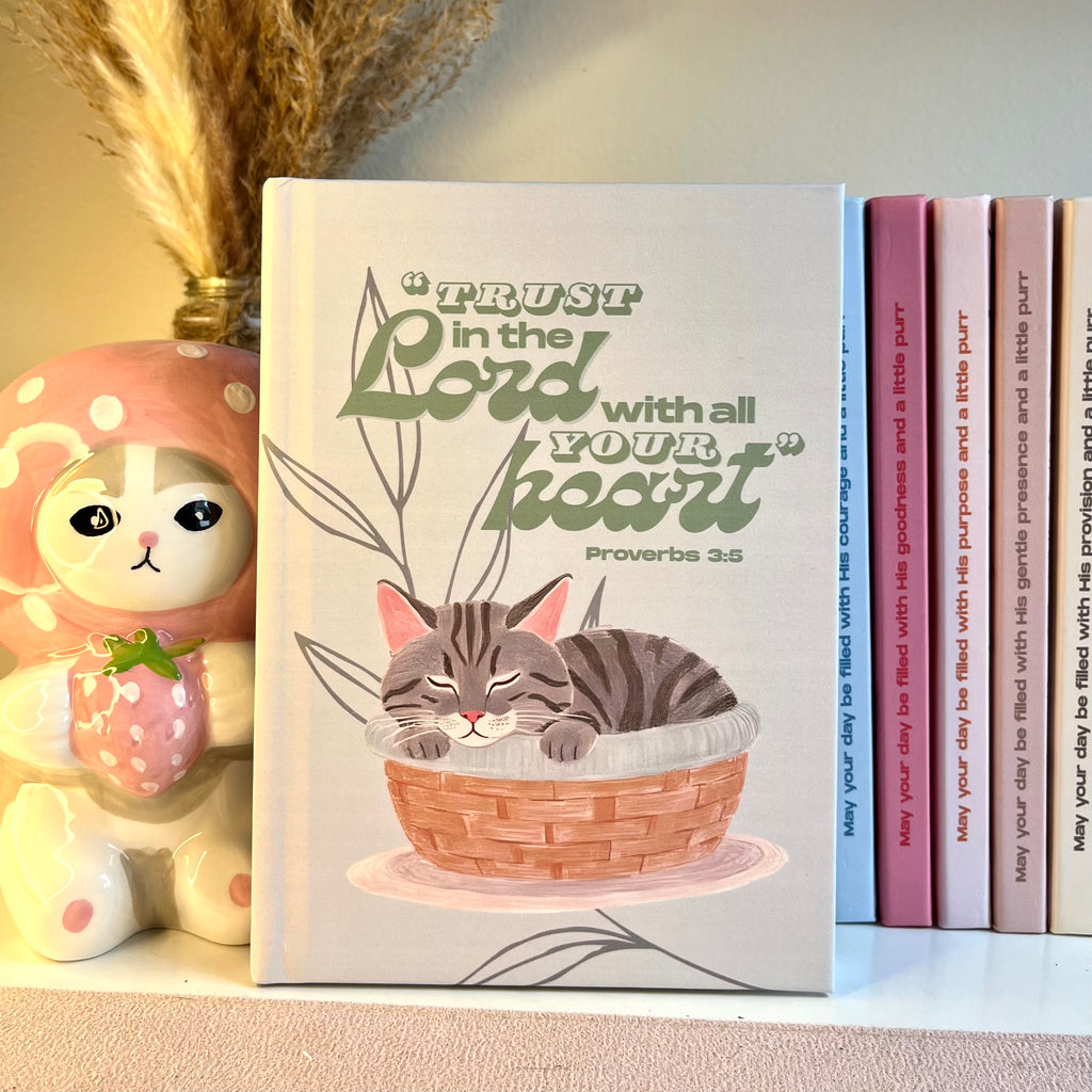 Notebook with a gray tabby cat illustration and motivational Bible quote on a shelf with decorative items.