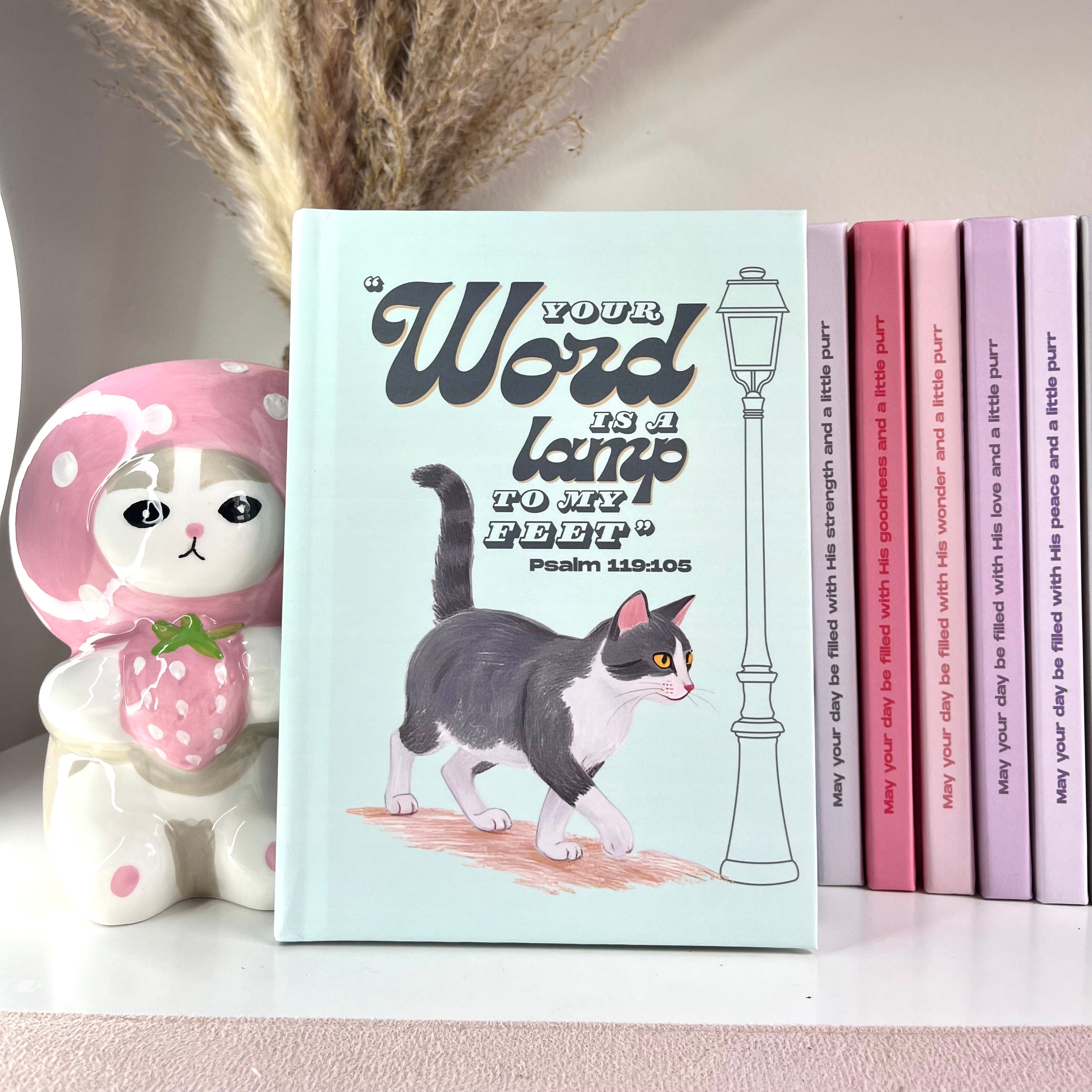 Mint color notebook with a gray tuxedo cat illustration and a Bible verse text on a shelf with decorative items