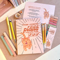 Notebook with orange cat illustration, pens, and stationery items on a textured surface