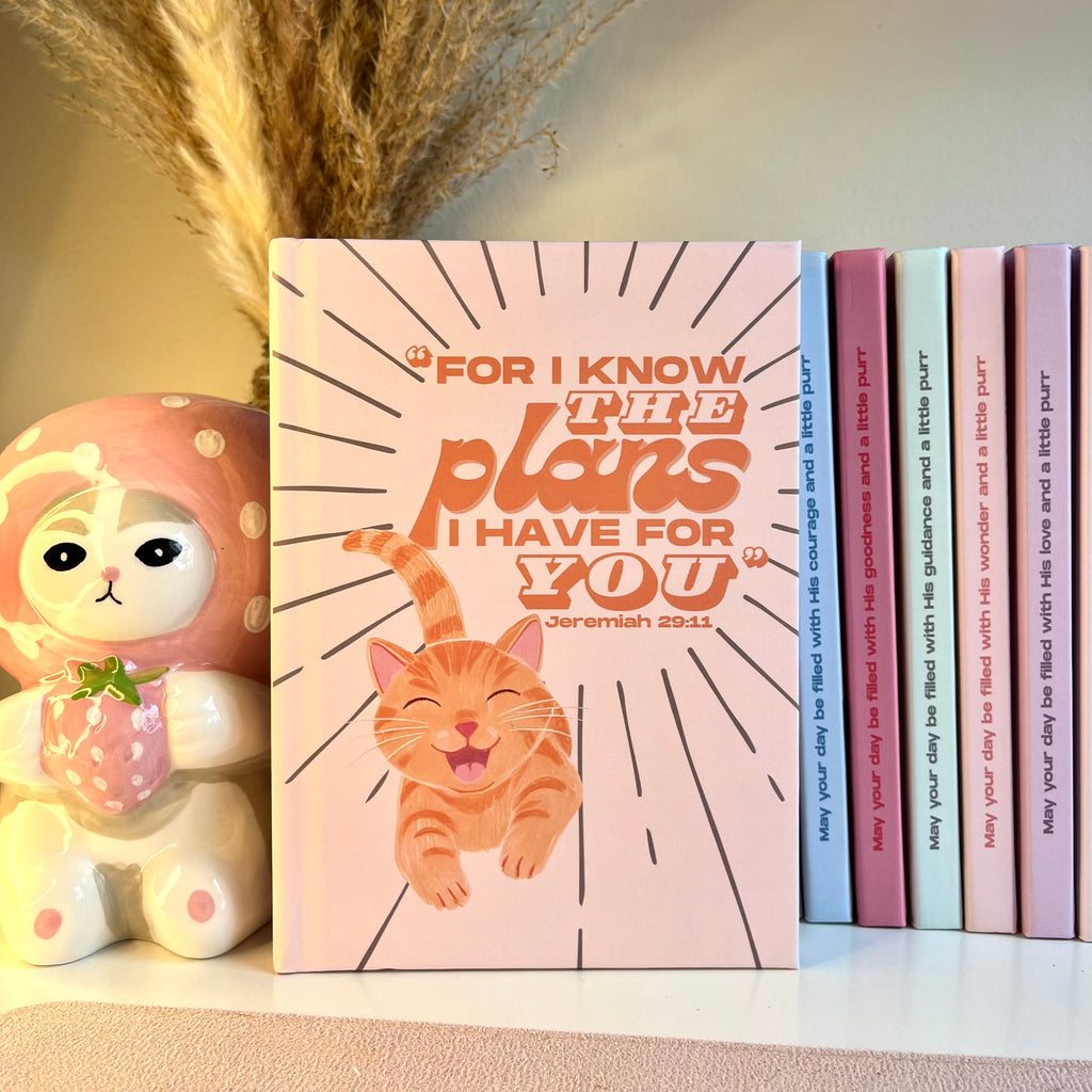 Orange notebook with an orange tabby cat illustration and motivational Bible quote, surrounded by decorative items.