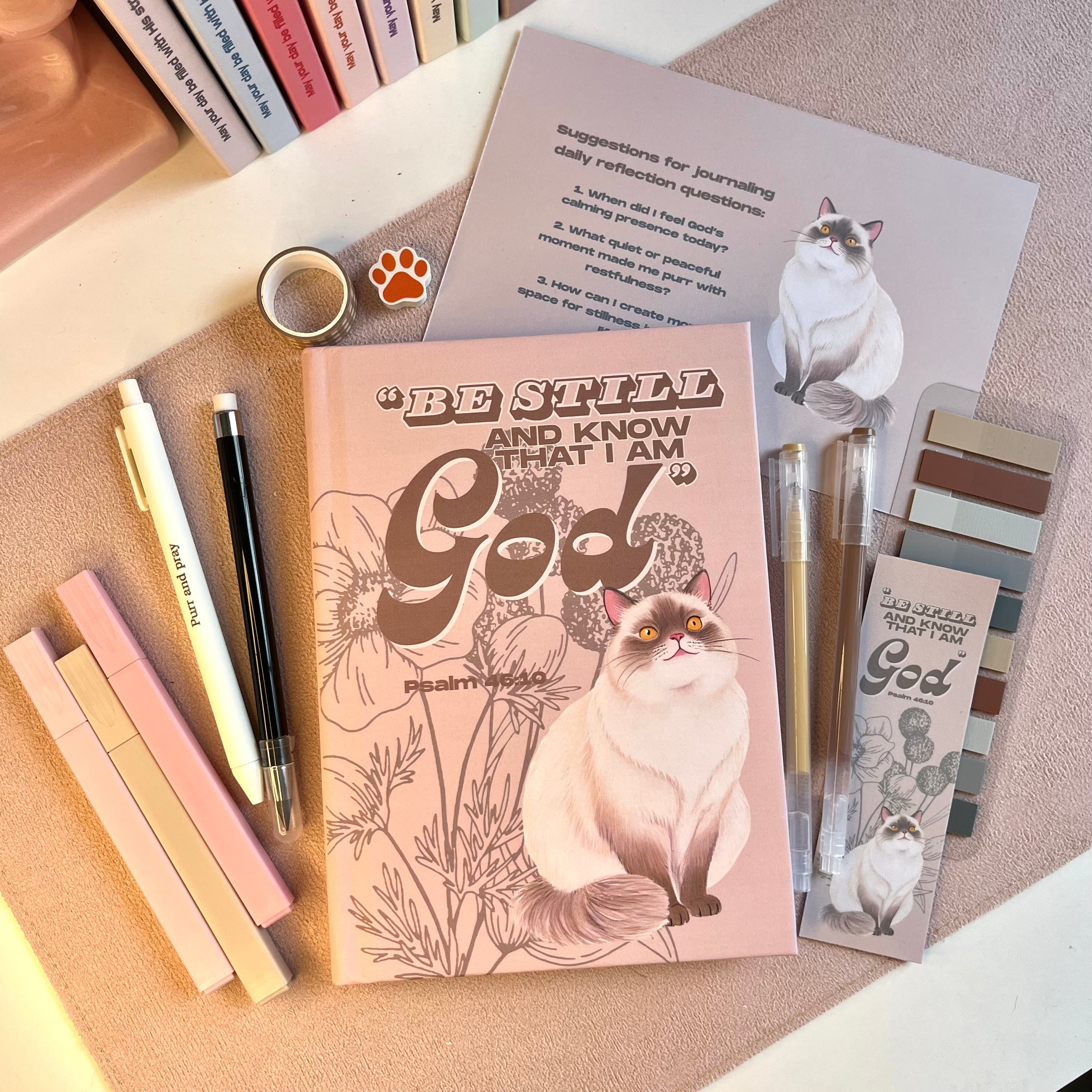 Notebook with cat illustration and 'Be Still and Know That I Am God' text, surrounded by pens and stationery items.