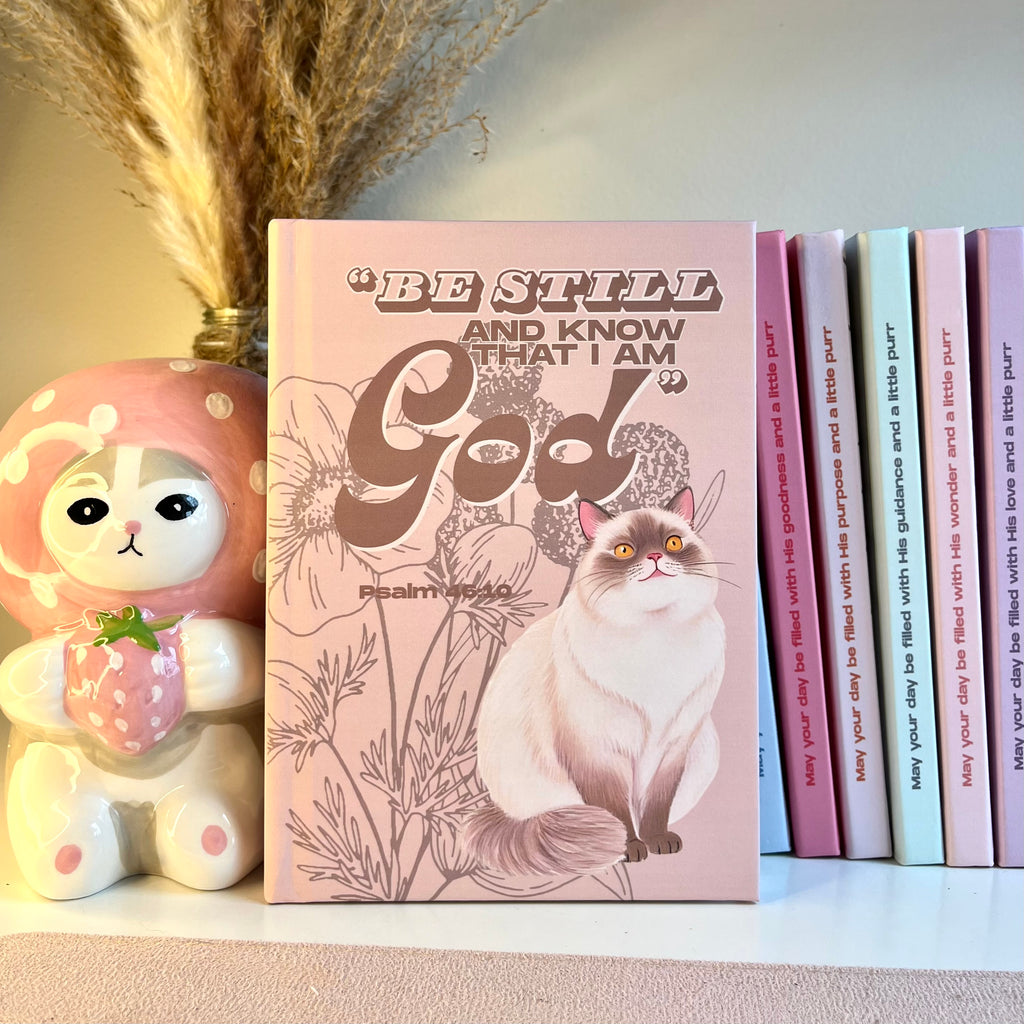 A beige notebook with a siamese cat illustration and motivational Bible quote, placed on a surface with decorative items.