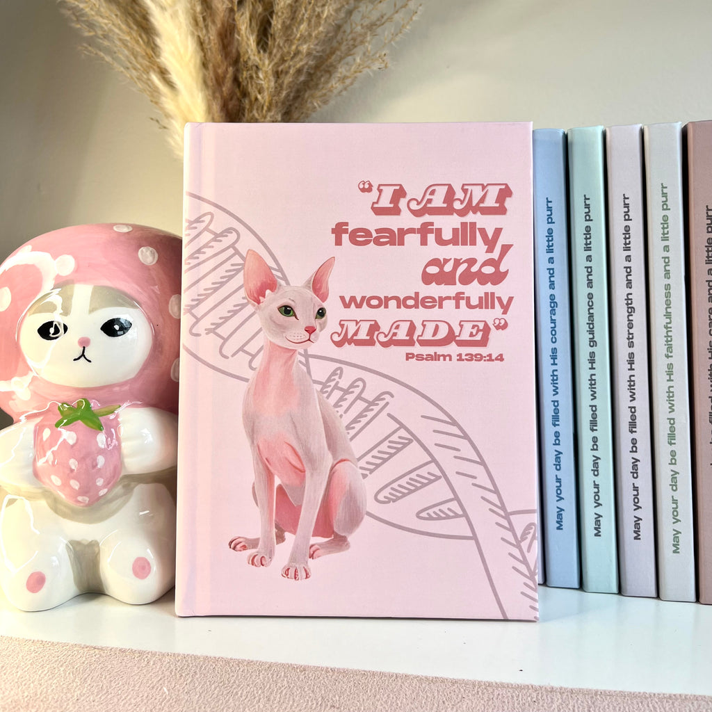 Pink notebook with a sphynx cat illustration and  Bible verse text, next to a decorative figurine and books.