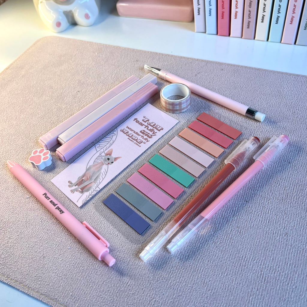 Pink stationery items including pens, markers, and sticky notes on a textured surface with books in the background.