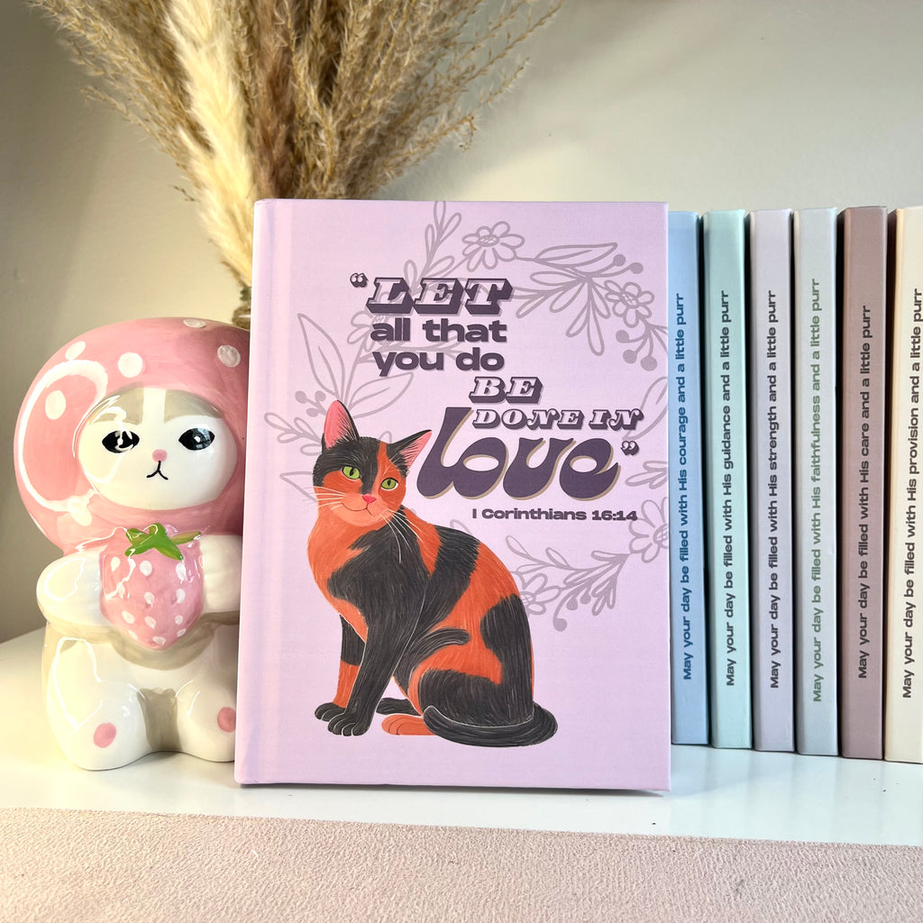 Purple notebook with a tortosieshell cat illustration and motivational quote from Bible on a shelf with decorative items.