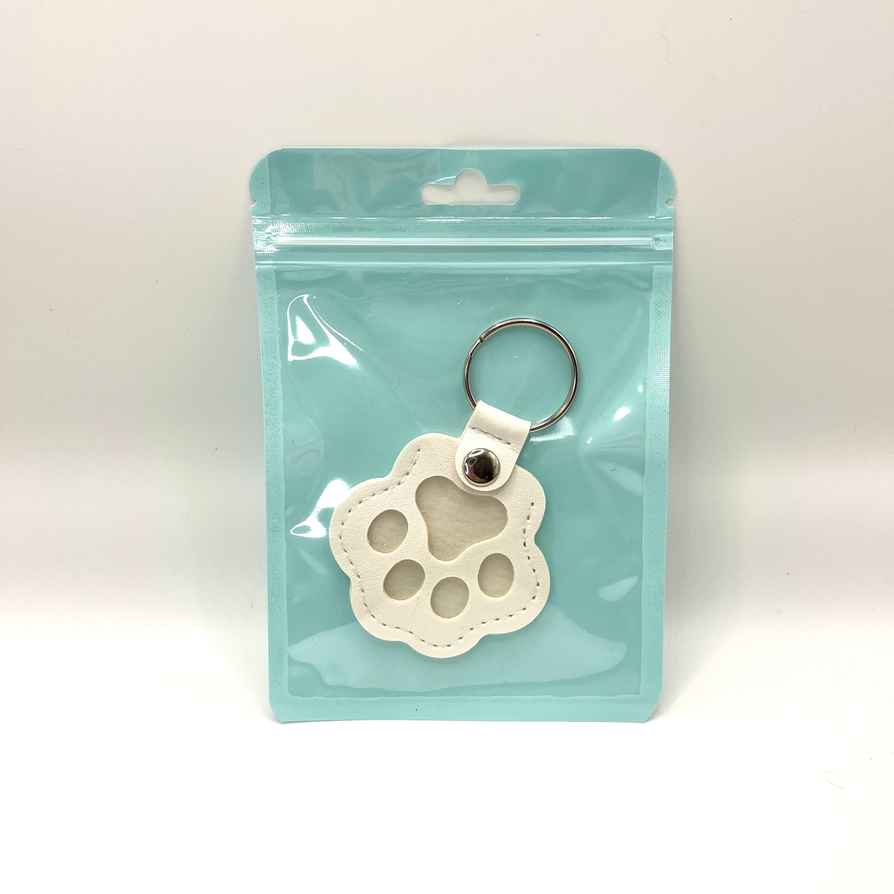 White faux leather paw keepsake keychain with mini fur vial displayed inside a plastic bag.
