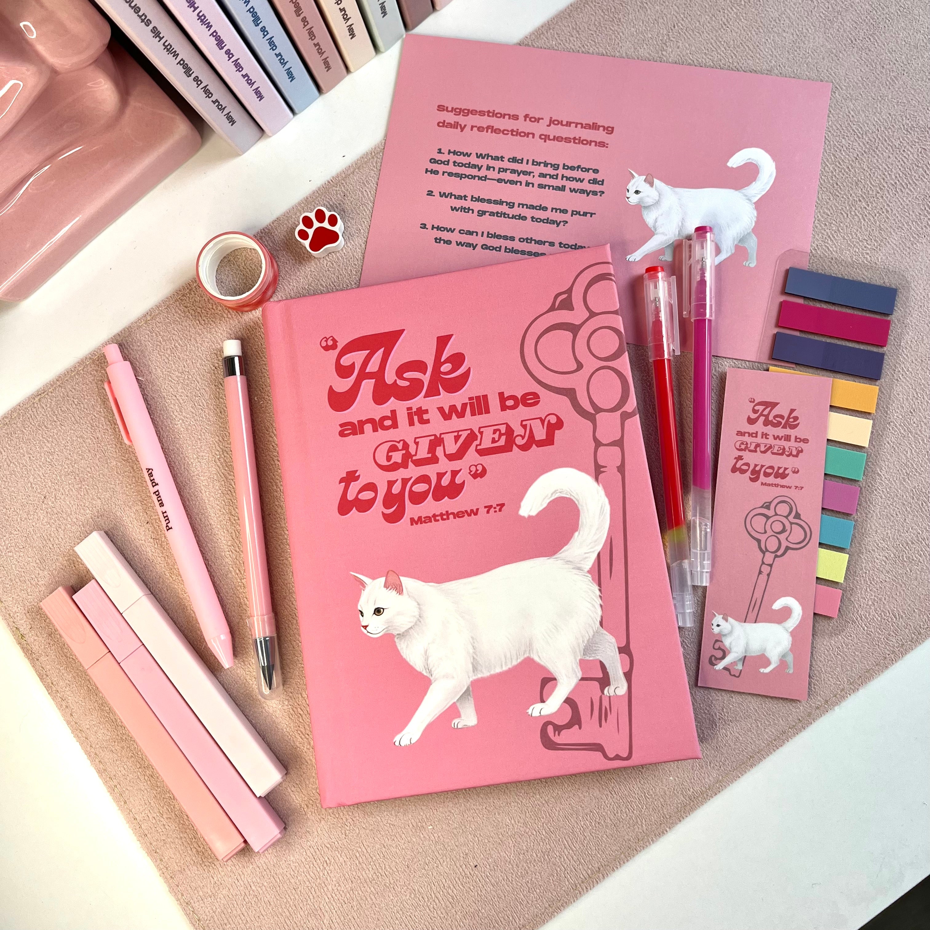 Pink notebook with white cat illustration and motivational quote, surrounded by pens and bookmarks on a textured surface.