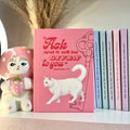 Pink notebook with a white cat illustration and motivational Bible quote, surrounded by decorative items.