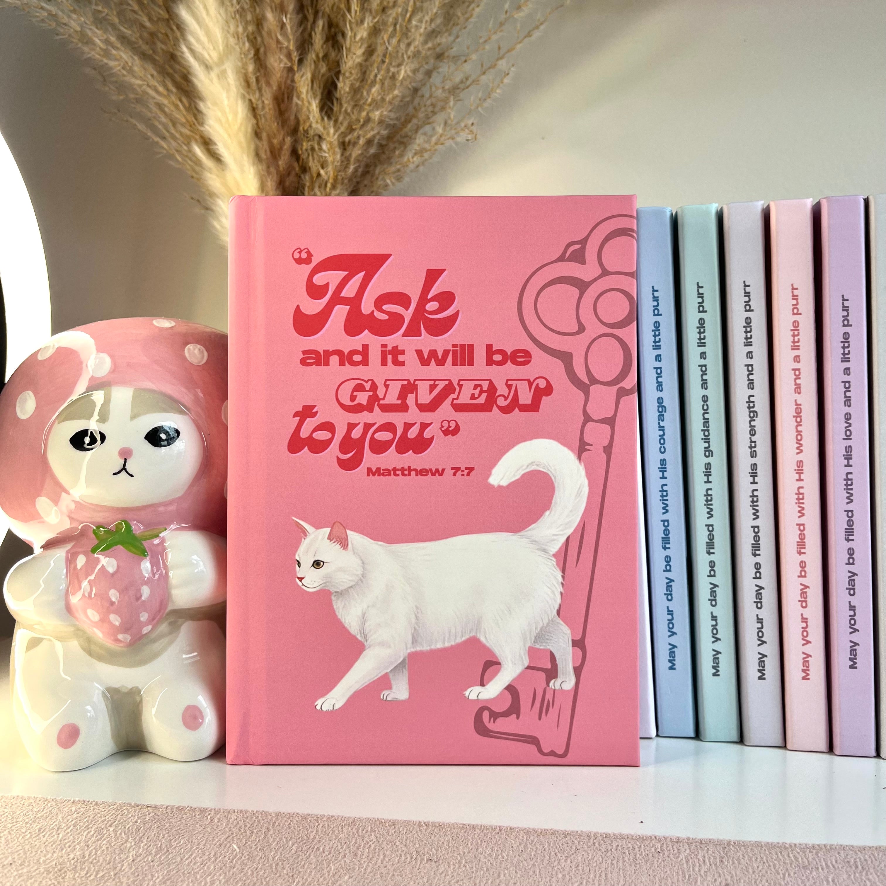 Pink notebook with a white cat illustration and motivational Bible quote, surrounded by decorative items.