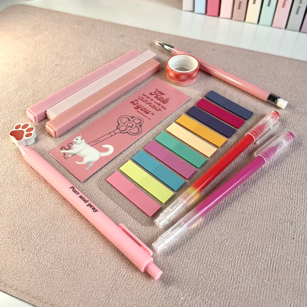 Set of pink stationery items including pens, sticky notes, and a small notebook with white cat illustration on a textured surface.