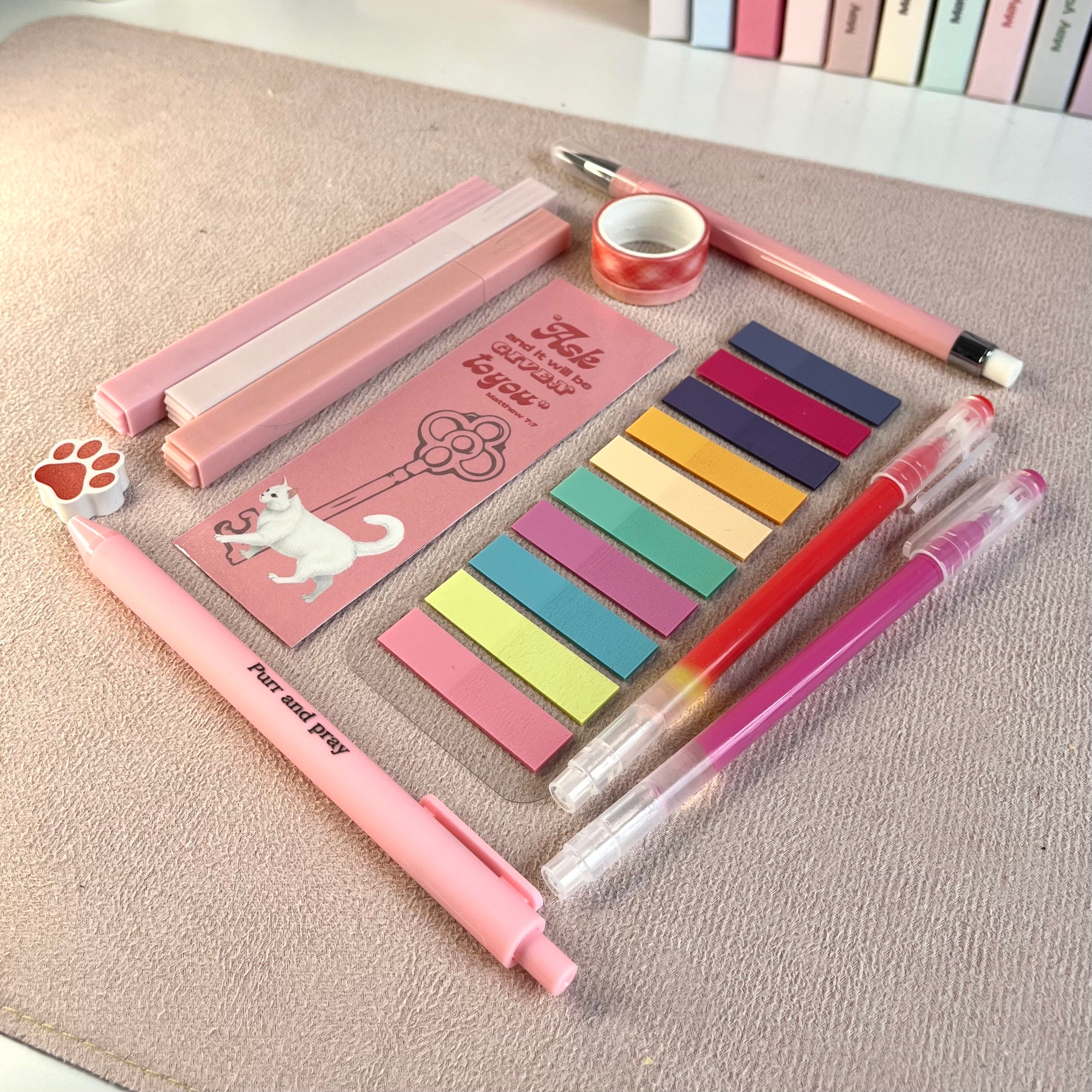 Set of pink stationery items including pens, sticky notes, and a small notebook with white cat illustration on a textured surface.